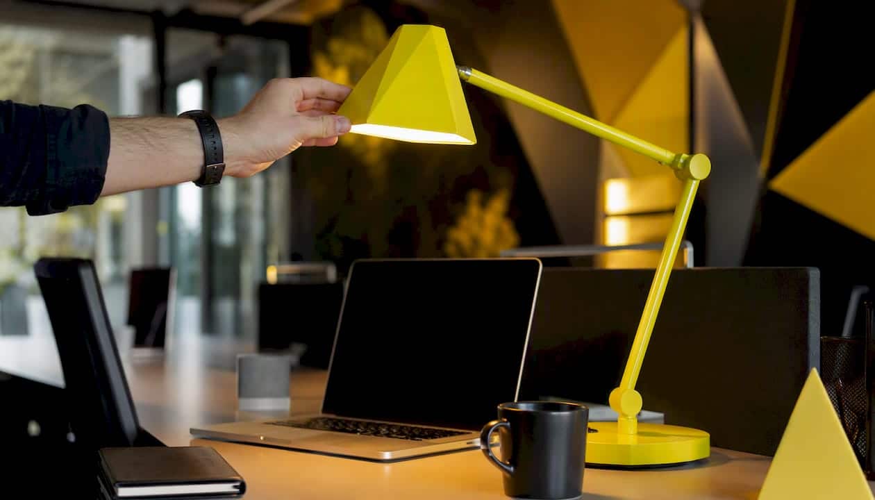 Une main ajuste une lampe de bureau jaune fluo posée sur la table d’une agence.